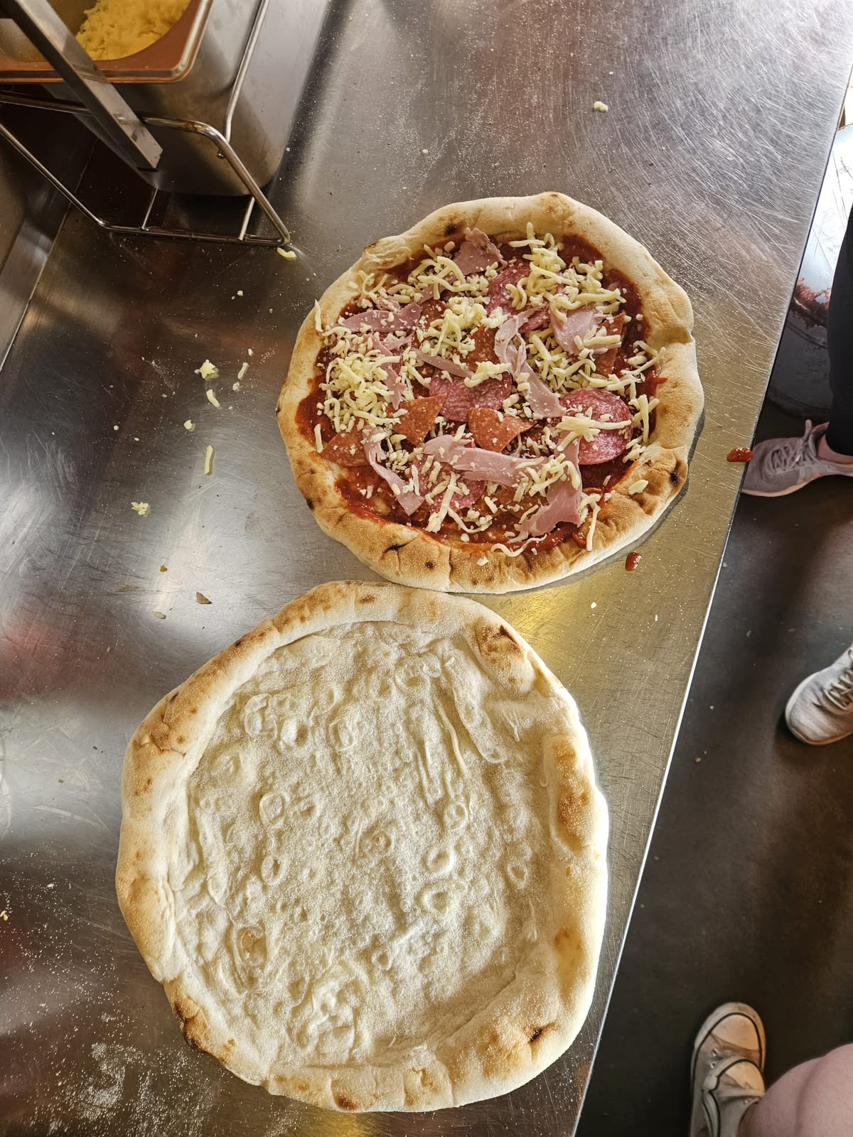 A plain base beside a topped pizza during kitchen preparation